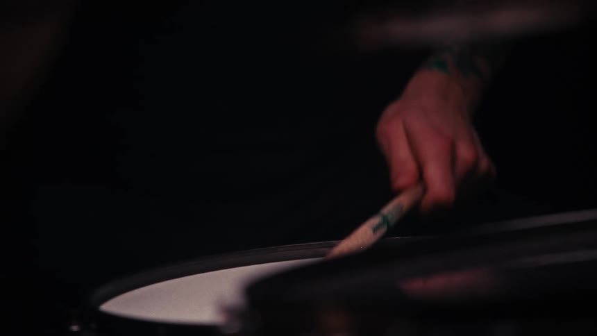 A closeup image shows a persons hands playing a drum with drumsticks with one hand wearing a brown bracelet