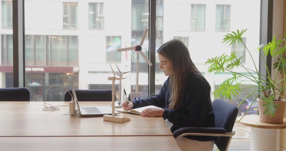 Young woman sitting at an office desk taking notes in a notebook facing an open laptop