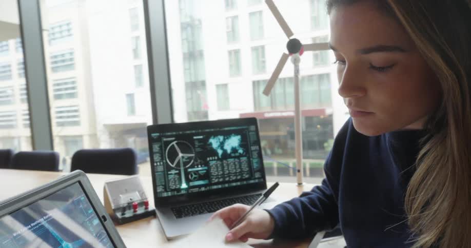 Young woman works in a bright room analyzing data on laptops and models of wind turbines