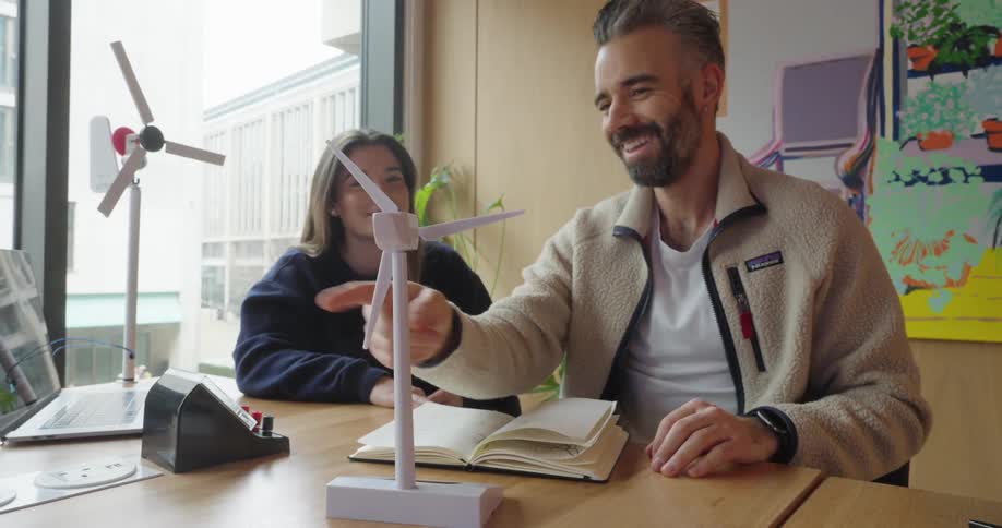 Man and a woman are sitting at a table in a modern setting surrounded by study materials and models of wind turbines