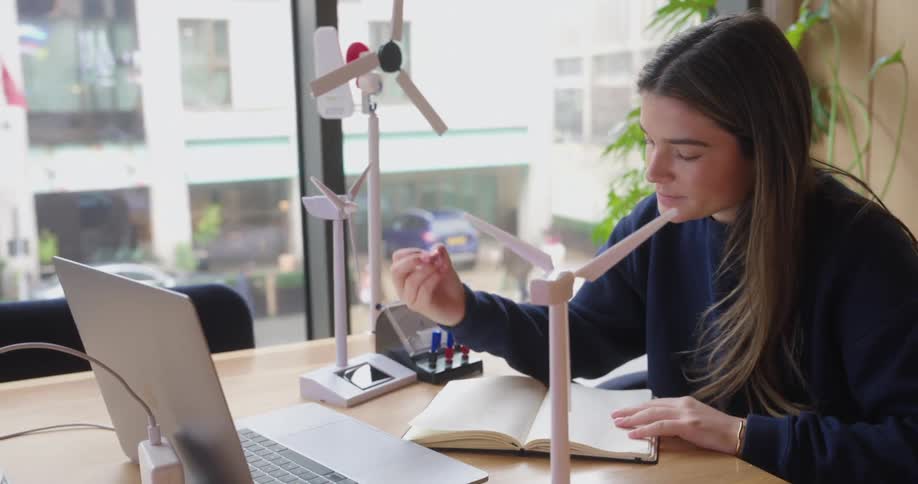 A young woman sits at a desk thoughtfully examining and then writing about model wind turbines with a laptop nearby