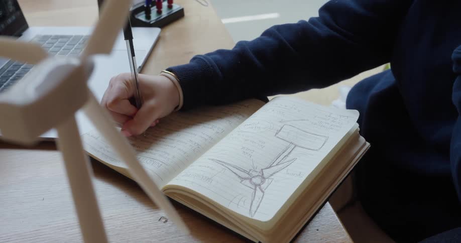 Person sketches a detailed drawing of a wind turbine in a notebook while holding a pen