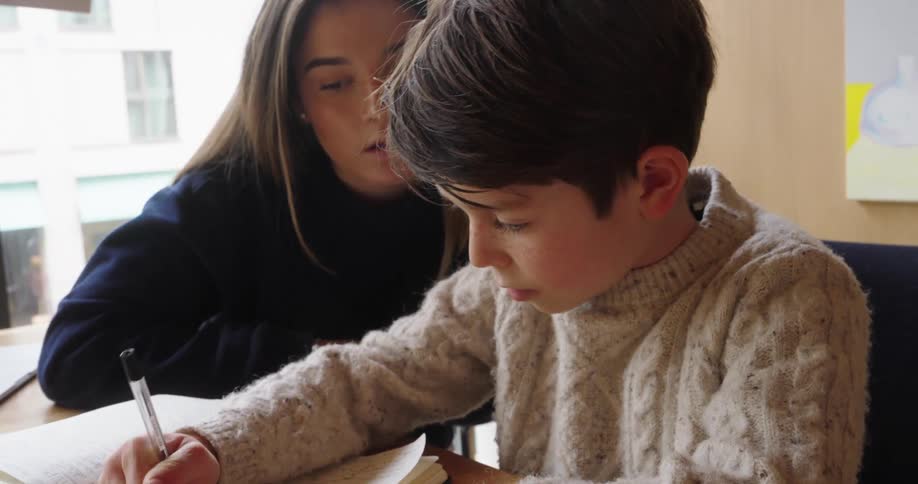 A teenage girl and helping a little boy while he is writing on a paper and they both are sitting