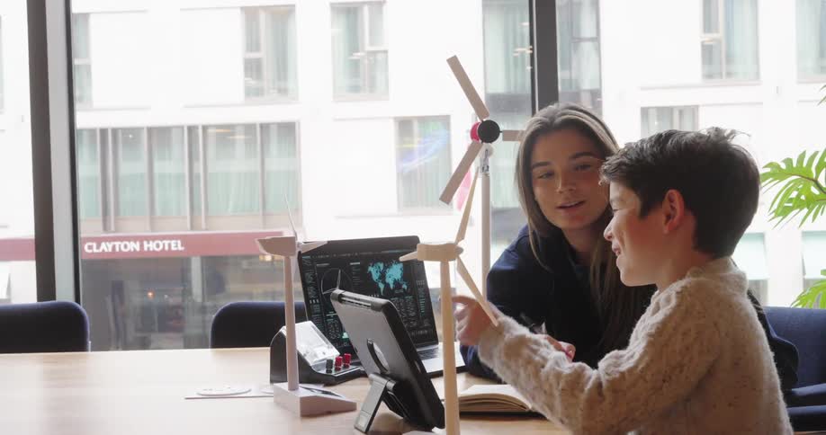 A woman and a young boy are seated at a table interacting with and discussing model wind turbines and a laptop displaying data