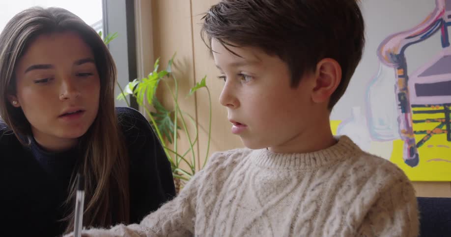 A young boy focused on writing sits at a table while a woman attentively watches him offering silent support