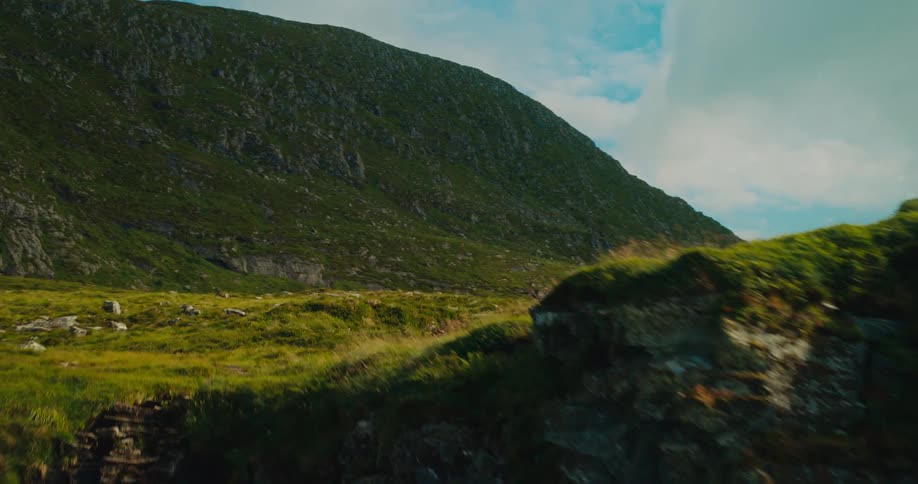 From an aerial perspective the camera rapidly flies towards a mountain cliff unveiling a scenic mountain pasture bathed in daylight capturing the vastness of the mountainous landscape