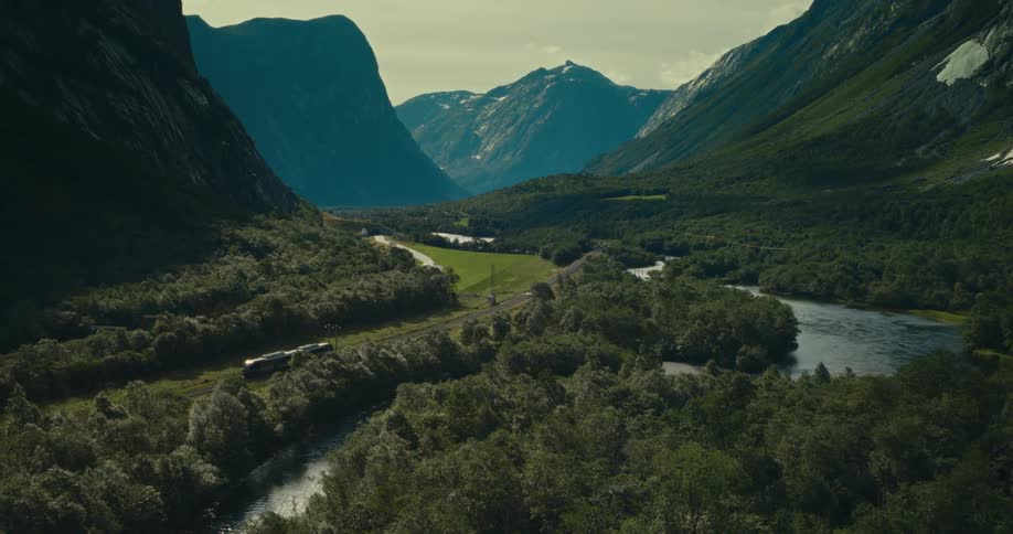 A train moves through a lush green valley surrounded by towering mountains and a winding river creating a serene and picturesque landscape