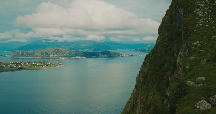 A scenic view from a grassy mountain slope overlooking a calm body of water with islands and distant mountains under a partly cloudy sky