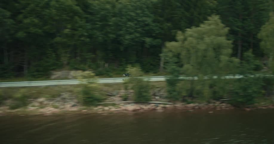 Motorcyclist rides along a waterfront highway passing a forested mountain on an overcast day