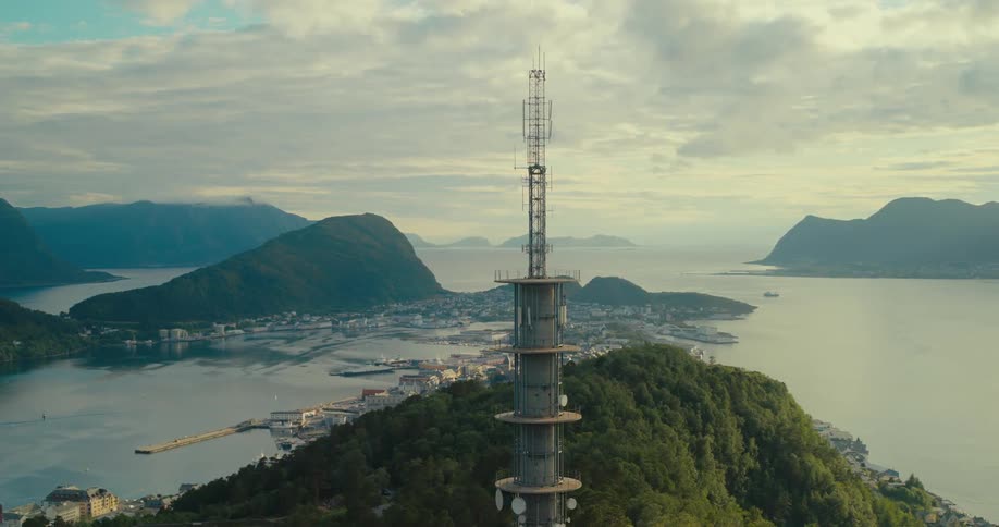 Telecommunication tower stands on a forested mountain near a coastal town on a foggy day