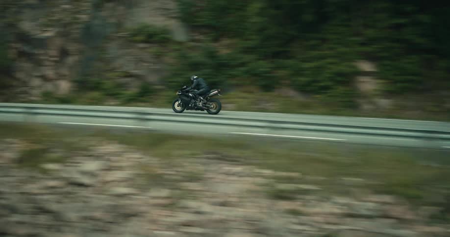 A motorcyclist in black gear rides a black sport bike along a road with a guardrail next to a forested area and a body of water