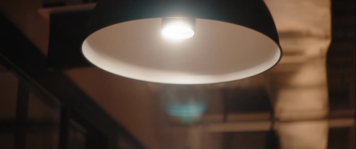 A dimly lit hallway with glass walls is illuminated by a row of ceiling lamps that are turning on casting a soft glow and highlighting the misty atmosphere in the distance
