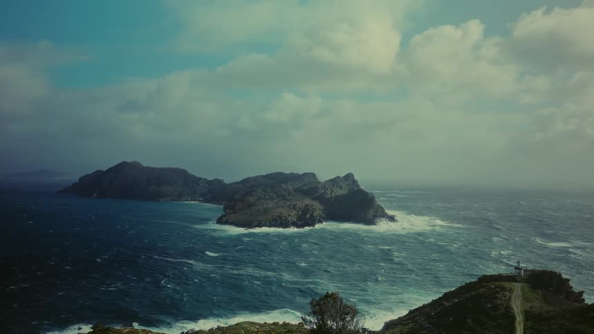 Aerial view of a rocky island in a choppy sea with a partial view of a coastline in the foreground