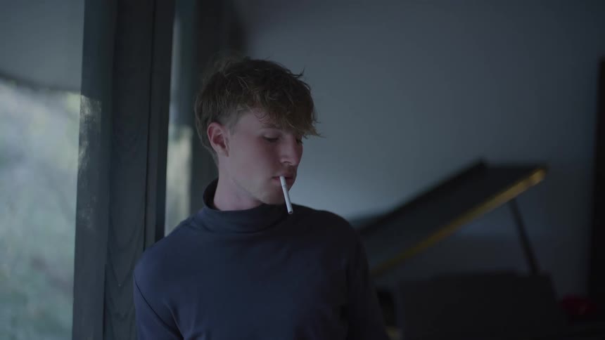 Young man with messy hair wearing a dark turtleneck looks thoughtfully out the window with a cigarette in his mouth