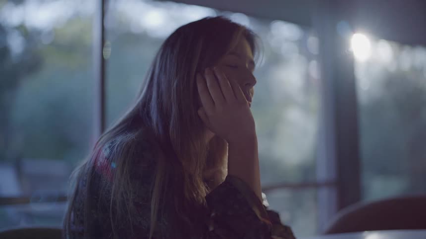 The face of a young woman resting her head on her hand and she is giving long blinks in an indoor environment