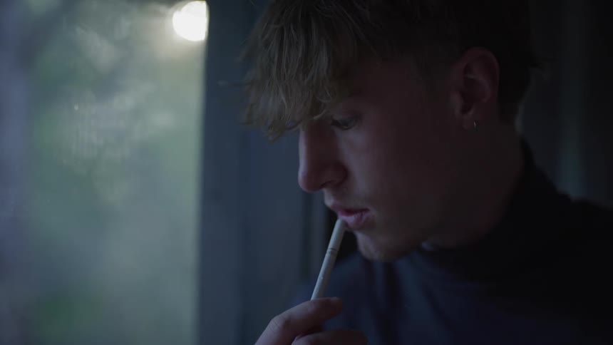A man is holding a cigarette by the window