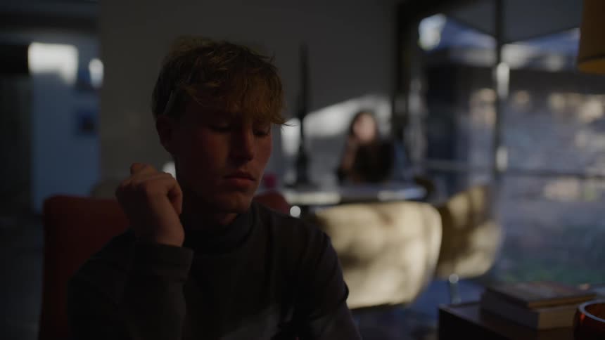 Blurred man in foreground while woman sitting at table looks thoughtful