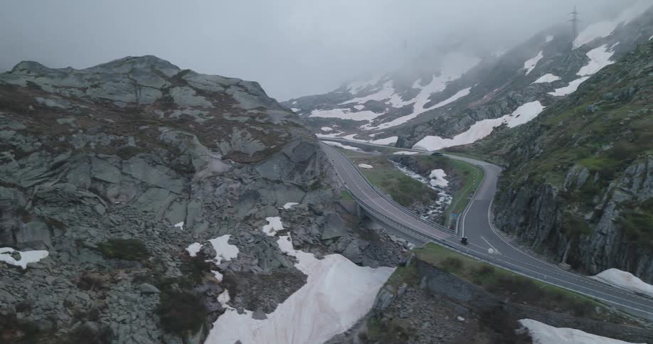 An aerial view of an asphalt road with a car passing by and it is located on a mountain range with snow on the ground