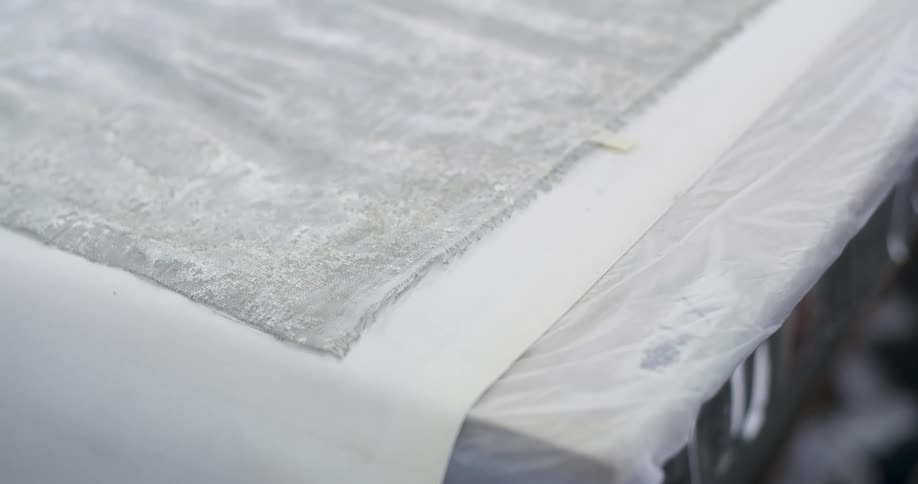 A piece of textured fabric rests on a white surface partially covered by a plastic sheet in a workspace hinting at an inprogress creative or craft endeavor