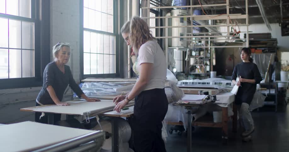 Three women work in a studio with two of them discussing a piece of fabric spread out on a table while the third watches in the background