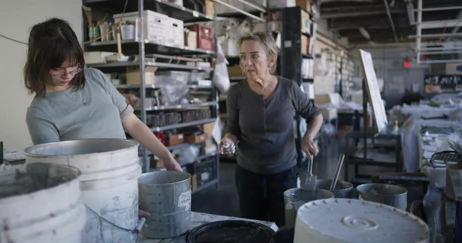 Inside a workshop during daytime the older woman while cleaning a tool and returning paint to a bucket is pictured talking to the younger woman who is intently inspecting a pail nearby