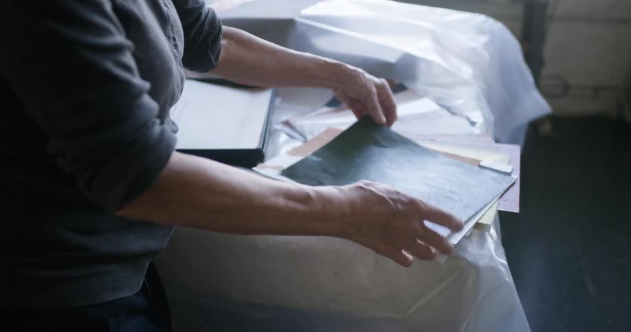 An elderly woman is going through some paint samples on a table top covered by plastic foil