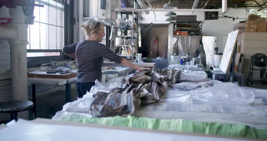 Woman arranges a large piece of patterned fabric on a work table in a studio lit by large windows