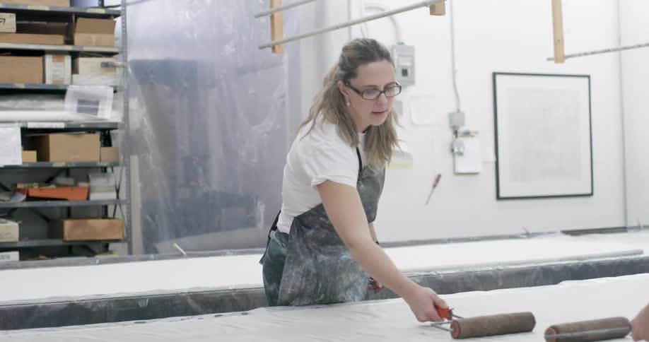 A woman wearing glasses and a dirty gray jumpsuit is using a brown paintbrush to brush the white surface of a table in front of her