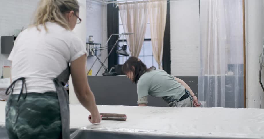 Two women work in an art studio using rollers to apply paint to a large surface