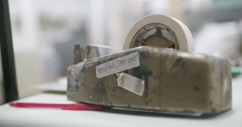 A paintsplattered tape dispenser labeled masking tape only rests on a busy workspace hinting at active craft or construction projects in progress