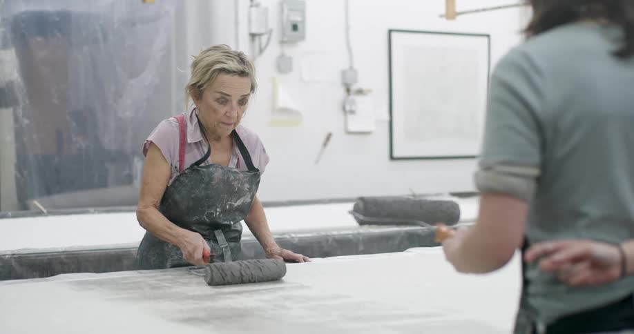 Two women paint a flat panel on a long worktable while working in a workshop