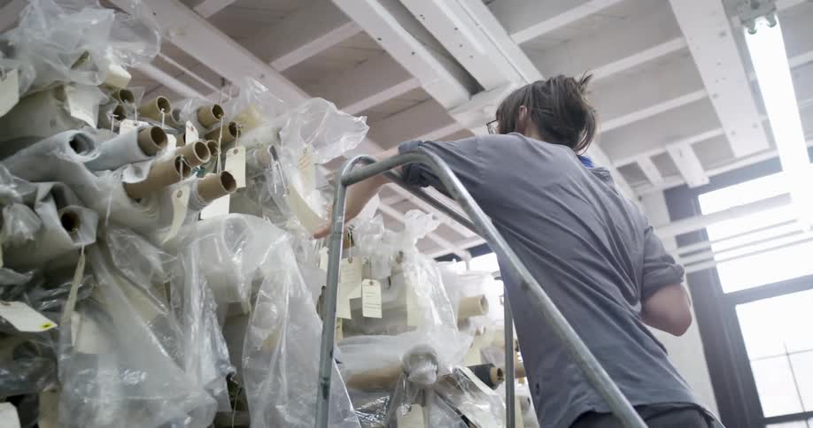 Woman climbs a ladder to reach rolls of fabric stored on high shelves inside a studio