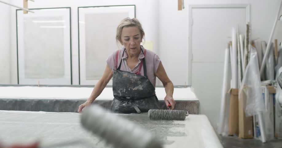 Woman wearing an apron applies paint with a roller to a large surface in an art studio