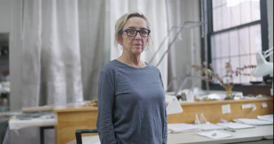 In a bright studio filled with papers and art supplies a woman with short blonde hair blue glasses and a striped shirt turns her head to the camera with a thoughtful expression