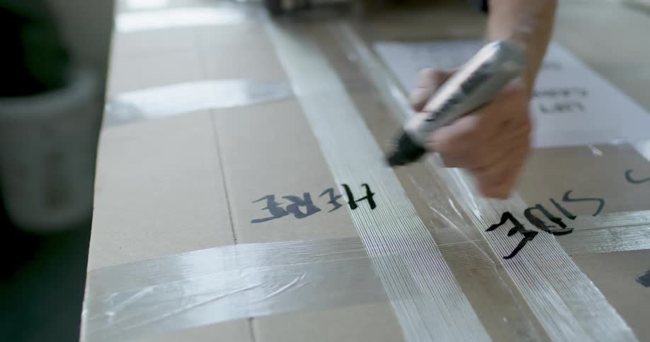 A persons hand writing on the surface of a cardboard box using a black marker
