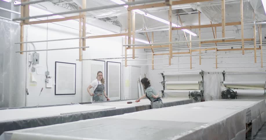 Two women in a factory using a roller and ironing on a table with fabrics