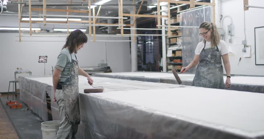 Women apply paint onto fabric while working in a large workshop