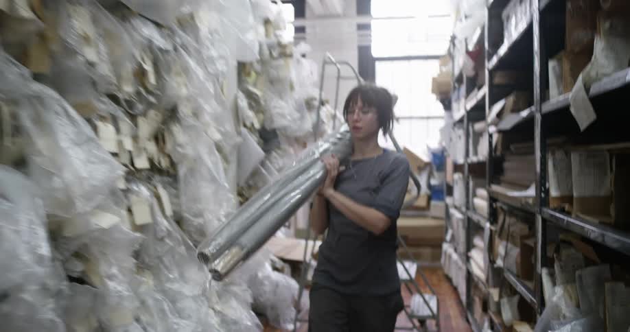 A woman wearing glasses and a darkcolored shirt carries a large rolled up material wrapped in plastic through a cluttered storage room filled with shelves and plastic wrapped items
