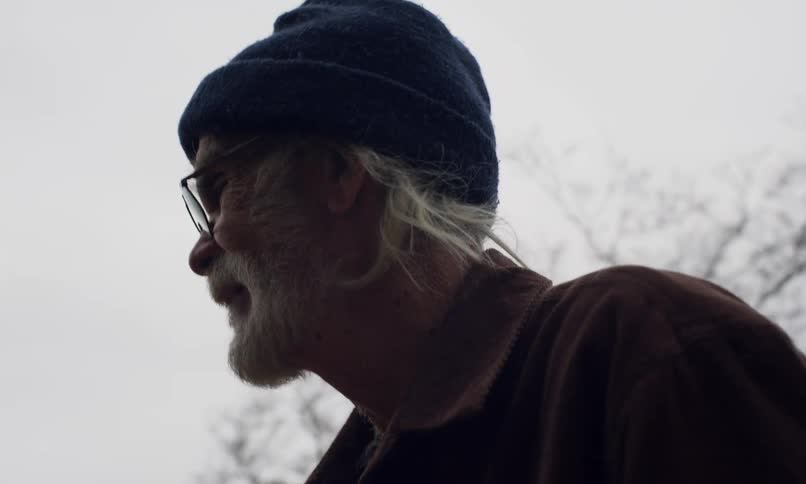 Bearded white haired man stands outdoors near a leafless tree