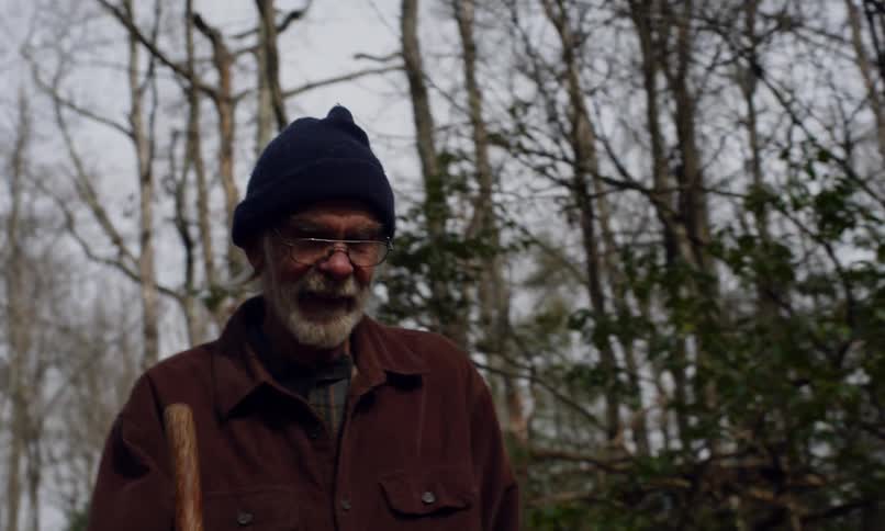 A man with a white beard wearing a blue beanie and a brown jacket is walking in a forested area with bare trees and some green foliage
