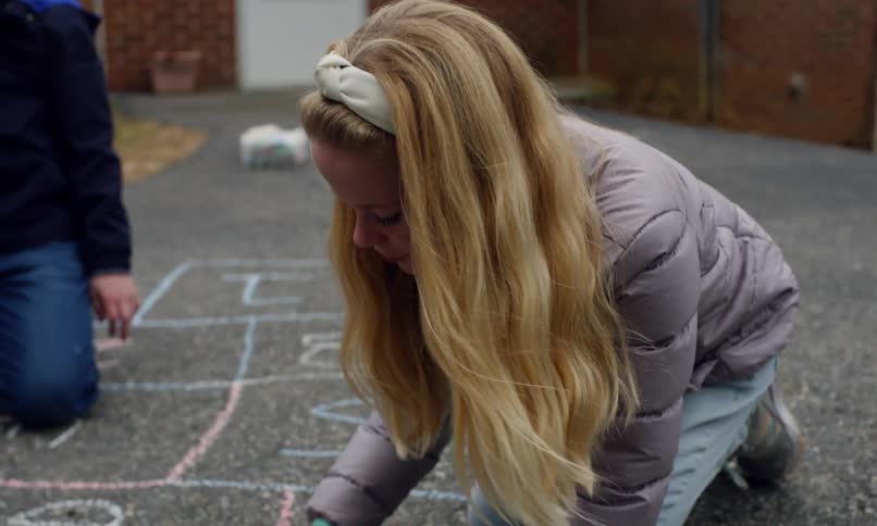 A young girl with long blonde hair wearing a lightcolored headband and a puffy jacket leans down as she carefully draws on the pavement with colorful chalk
