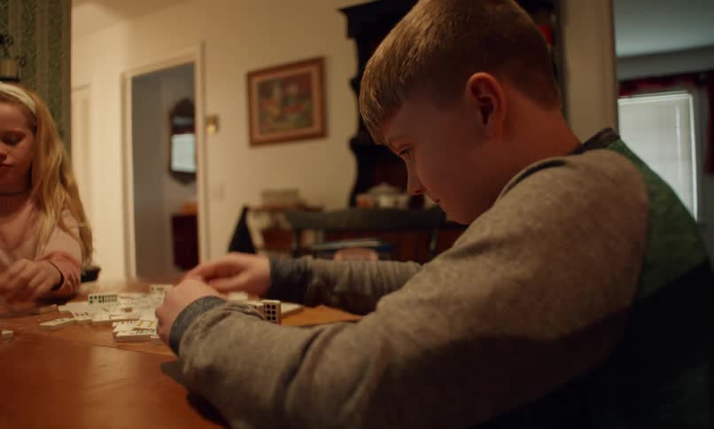 A young boy and girl are sitting at a wooden table engaged in a board game with dice and tiles in a warmly lit home setting