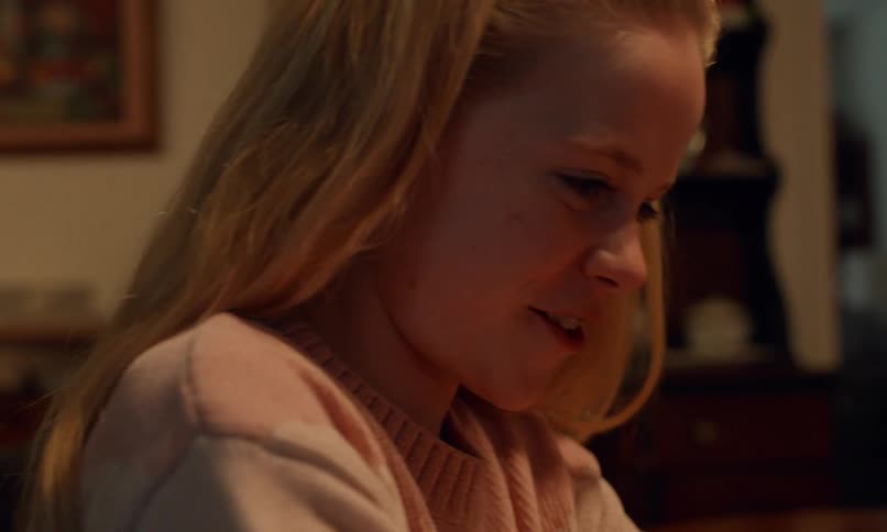 A blonde girl with a soft smile and a focused expression sits in a warmly lit room seemingly engaged in a heartfelt moment as the cozy background adds to the intimate atmosphere