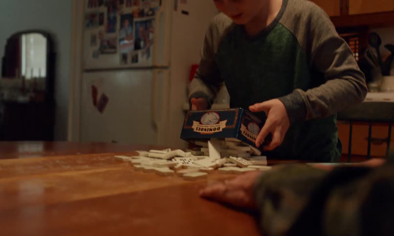 Boy dumps dominoes onto a wooden table while an elderly hand rests in front