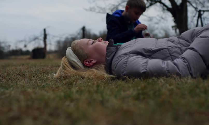 A young girl lies on the grass eyes closed while a boy in the background plays with a bubble wand