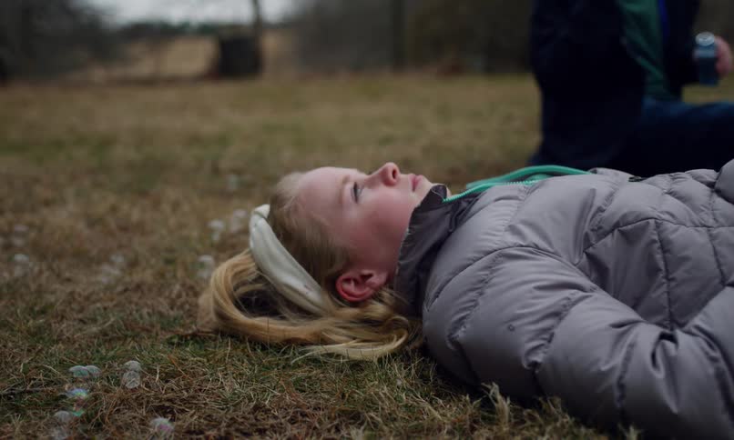 A young blonde girl lies still in a grassland while another child plays with bottled bubbles next to her