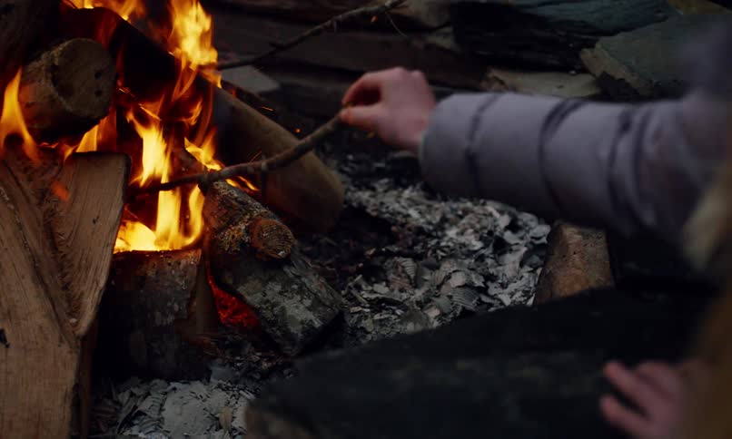 Person in a coat stirs a fire with a twig as the flames burn the wood