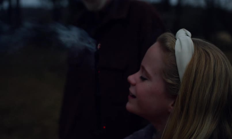 A girl blows on a stick with a small fire to extinguish it near an elderly man in the countryside at dusk