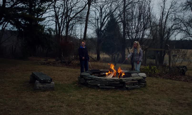 Two children burn the tips of sticks in a fire pit in the countryside at dusk