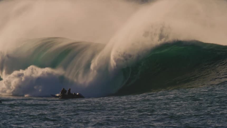 A surfer is riding a large powerful wave with a dramatic crest of white foam with other surfers visible in the background
